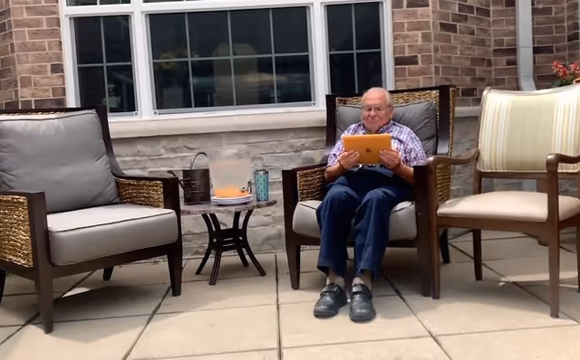 An elderly man sitting on a cushioned outdoor chair on a patio, holding a tablet. There are two other chairs nearby and a small round table with a pitcher and a glass on it. The background shows a brick wall with windows.