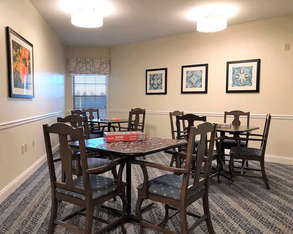 A well-lit room with multiple wooden tables and chairs arranged for seating. On the tables, there are puzzle boxes and partially completed puzzles. The room has carpeted flooring, light-colored walls with decorative molding, and framed artwork hanging on the walls. A window with blinds and a patterned valance is visible in the background.
