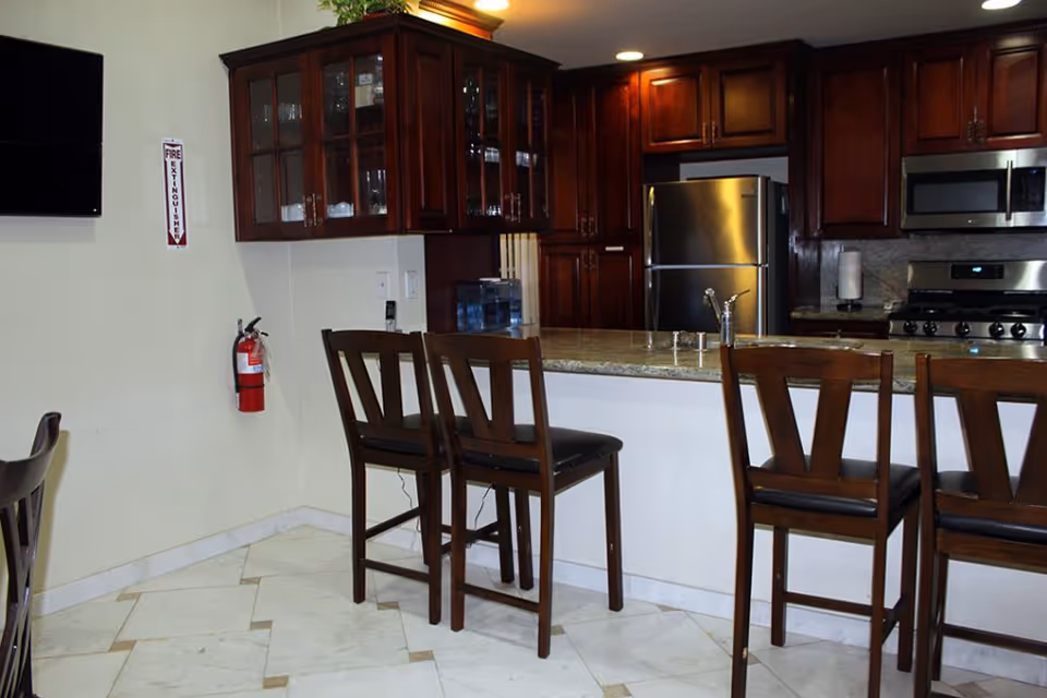 Interior view of a kitchen area with dark wooden cabinets, a stainless steel refrigerator, stove, and microwave. There is a granite countertop with four wooden bar stools. A fire extinguisher is mounted on the wall next to a flat-screen TV.