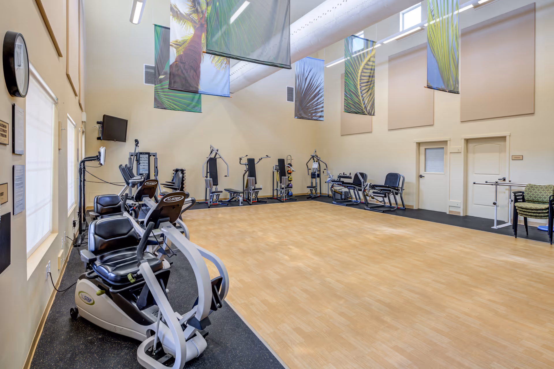 A spacious fitness room with exercise equipment including recumbent bikes and various strength training machines. The room has high ceilings with large windows near the top, decorative banners with palm leaf designs hanging from the ceiling, and light-colored wooden flooring in the center. There are chairs and a TV mounted on the wall.