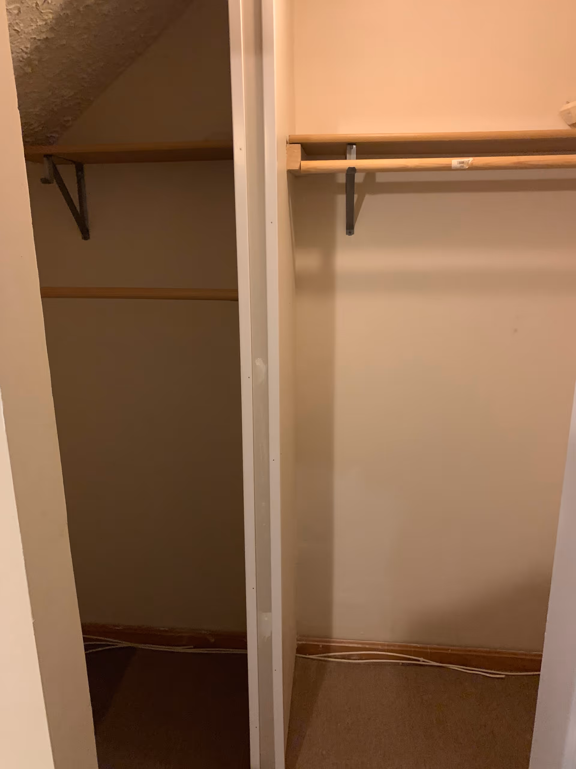 Interior view of a closet with two sections separated by a vertical divider. Each section has a wooden shelf and a hanging rod. The closet walls are beige, and the floor is carpeted. There is a visible cable running along the baseboard.
