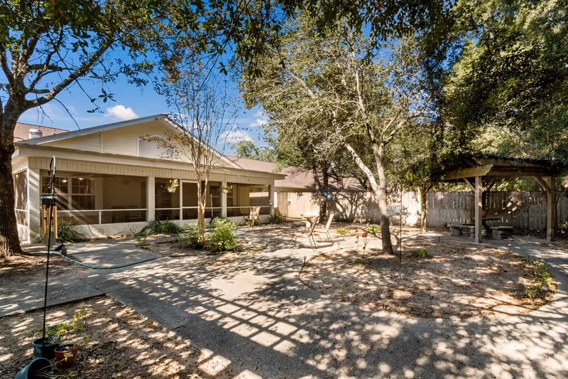 Outdoor courtyard area with a paved walkway, trees, and garden beds. There is a covered patio attached to a building on the left side, and a wooden pergola structure on the right side. Several chairs are placed around the courtyard, and the area is shaded by trees under a clear blue sky.
