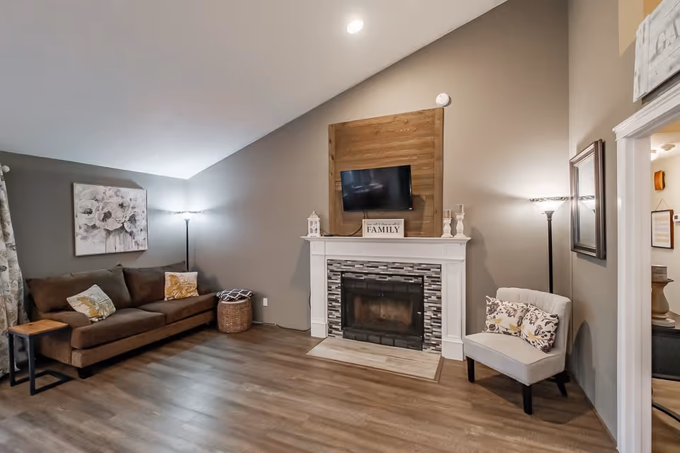 A cozy living room with a brown sofa adorned with decorative pillows on the left, a floor lamp behind it, and a floral painting on the wall. In the center is a fireplace with a tiled surround and a wooden panel above it holding a mounted flat-screen TV and decorative items including a sign that says 'Family'. On the right side, there is a light-colored armchair with patterned pillows, another floor lamp, and a doorway leading to another room.
