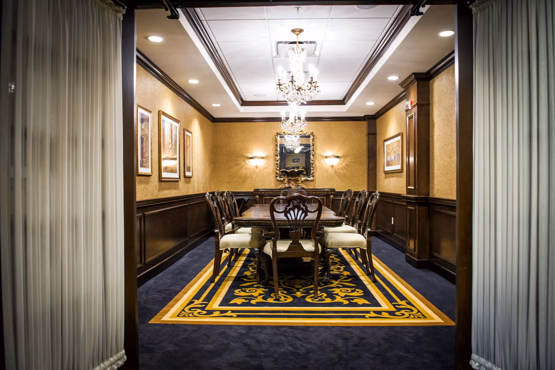 A formal dining room with a long wooden table surrounded by eight upholstered chairs. The room features dark wood paneling on the walls, a decorative blue and gold patterned carpet, framed artwork, a large ornate mirror, and two crystal chandeliers hanging from the ceiling.
