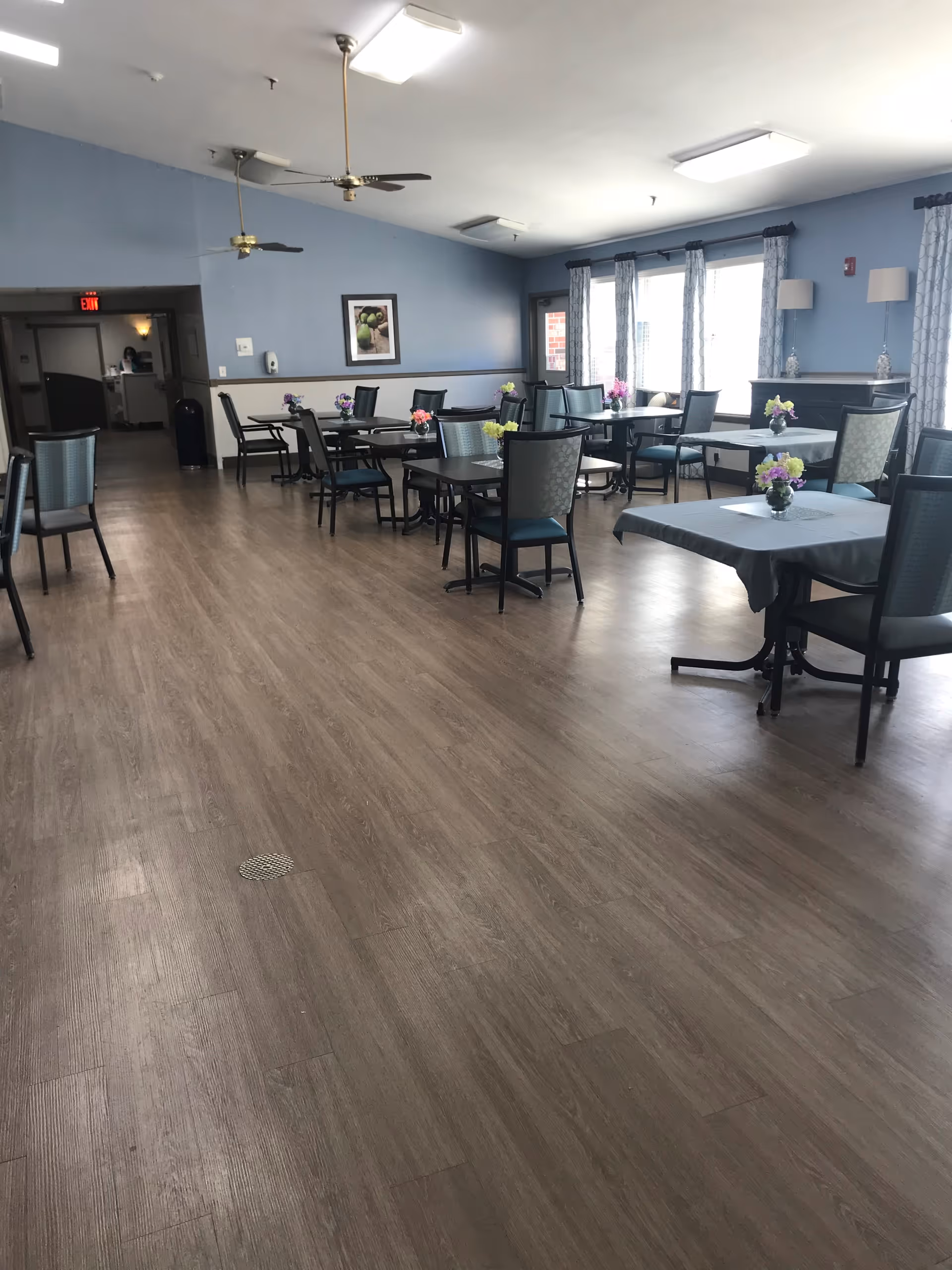 A spacious dining room with multiple tables and chairs arranged neatly. Each table has a small floral centerpiece. The room has light blue walls, large windows with curtains allowing natural light, ceiling fans, and wood flooring.