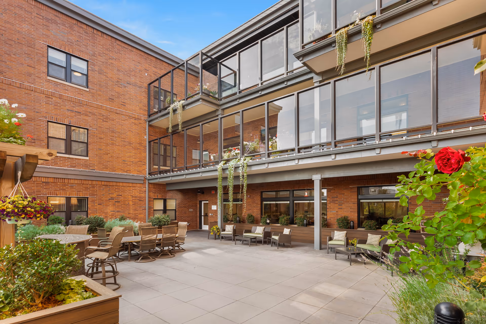 Outdoor patio area at Quail Park of West Seattle featuring multiple seating arrangements including tables with chairs and cushioned lounge chairs. The space is surrounded by brick walls with large windows and balconies adorned with hanging plants and flowers. The patio is decorated with various potted plants and flowers, creating a welcoming and relaxing environment under a clear blue sky.