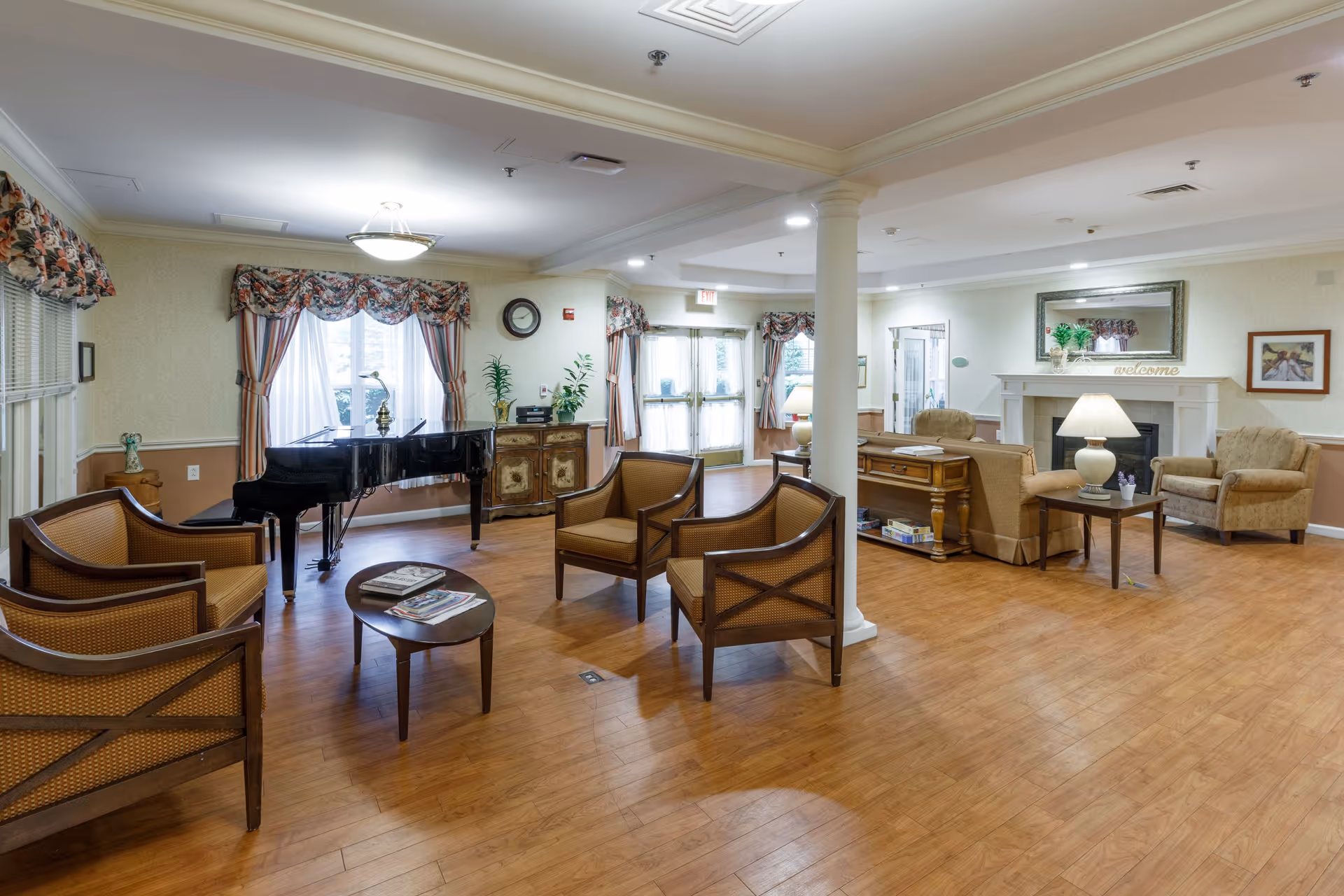 A spacious and well-lit assisted living facility common area with wooden flooring, featuring a black grand piano near windows with floral curtains, several wooden chairs with cushions arranged around a small oval table, a seating area with armchairs and a sofa around a fireplace with a 'welcome' sign, a large mirror above the fireplace, and a side table with a lamp.