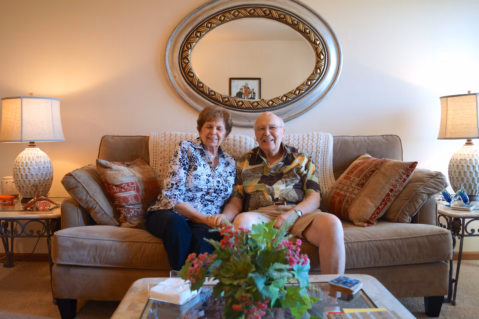 Two older adults sit on a couch in a warmly lit living room with decorative pillows, table lamps, a round wall mirror, and a coffee table with a floral arrangement.