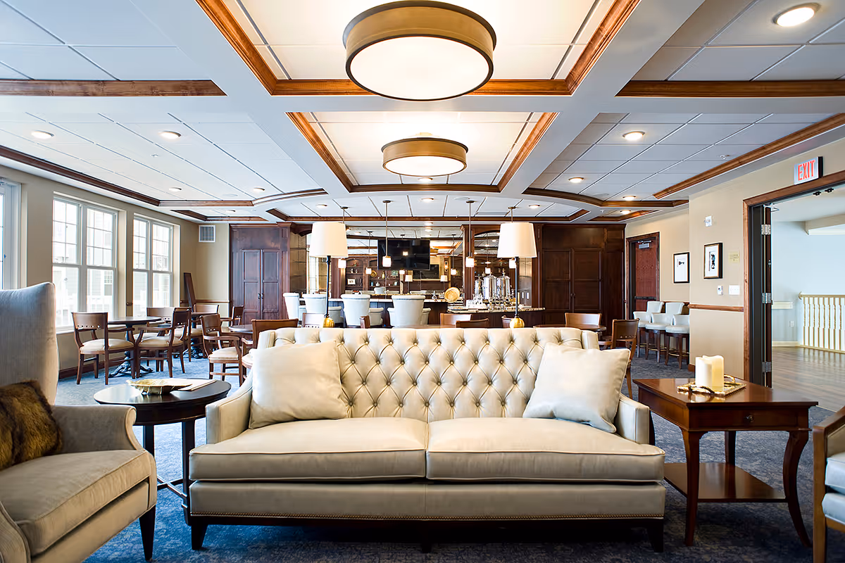 A spacious and well-lit common area in a senior living facility featuring a beige tufted sofa with two cushions in the foreground, flanked by armchairs and wooden side tables with decorative candles. In the background, there are multiple dining tables and chairs near large windows, a bar area with high stools, and elegant ceiling lights with wooden trim accents.