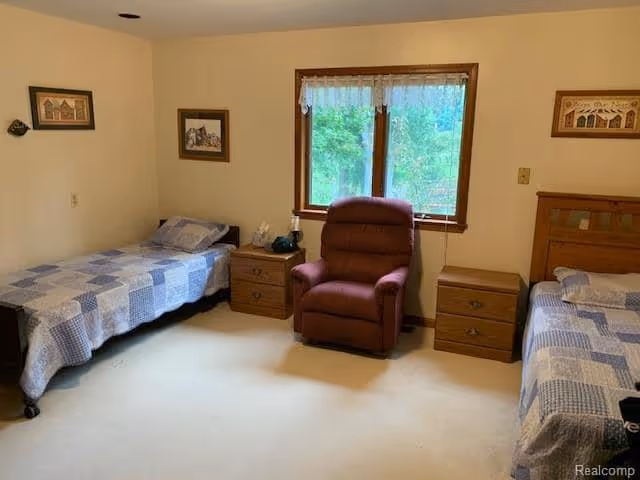 A simple bedroom with two single beds on opposite sides of the room, each with a blue and white patchwork quilt. Between the beds are two wooden nightstands, one with a lamp and a phone. A brown recliner chair is positioned in front of a window with white lace curtains, and the walls are decorated with framed pictures.