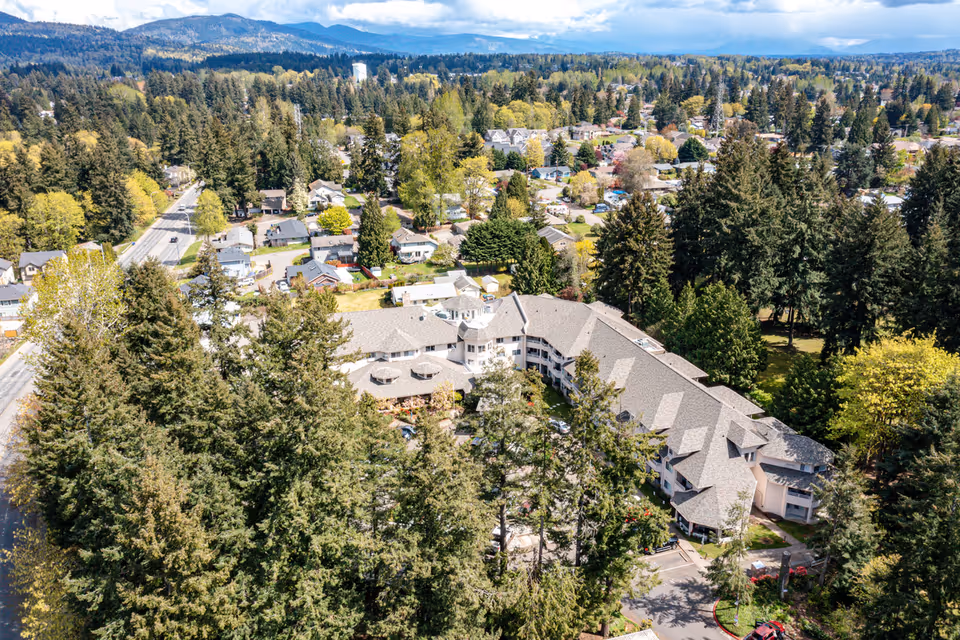 Aerial view of Solstice Senior Living at Renton surrounded by tall trees and residential houses, with mountains visible in the background under a partly cloudy sky.