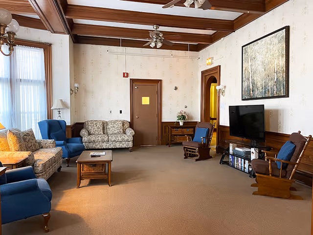 A spacious living room with vintage decor featuring patterned sofas, blue armchairs, wooden rocking chairs, a wooden coffee table, a flat-screen TV on a stand, and a large framed painting on the wall. The room has beige carpet, wallpaper with a subtle pattern, wooden ceiling beams, and large windows with sheer curtains allowing natural light to enter.