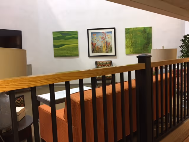 Interior view of a senior living community lounge area with an orange sofa, a patterned armchair, a side table with a lamp, and three colorful paintings on a white wall. A wooden railing with black vertical bars is in the foreground.