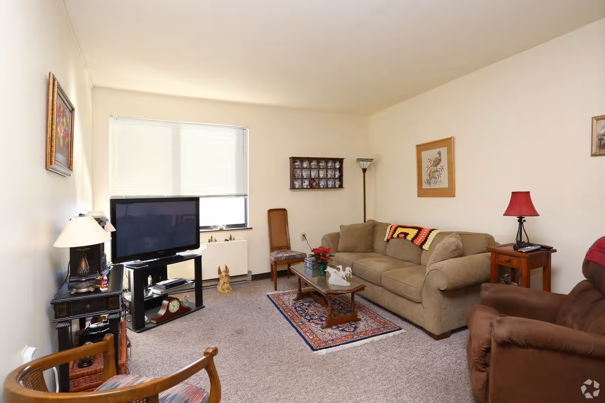 A cozy living room with a beige sofa, a brown recliner chair, a wooden coffee table on a small patterned rug, a TV on a stand, and various decorative items including framed pictures on the walls and table lamps.