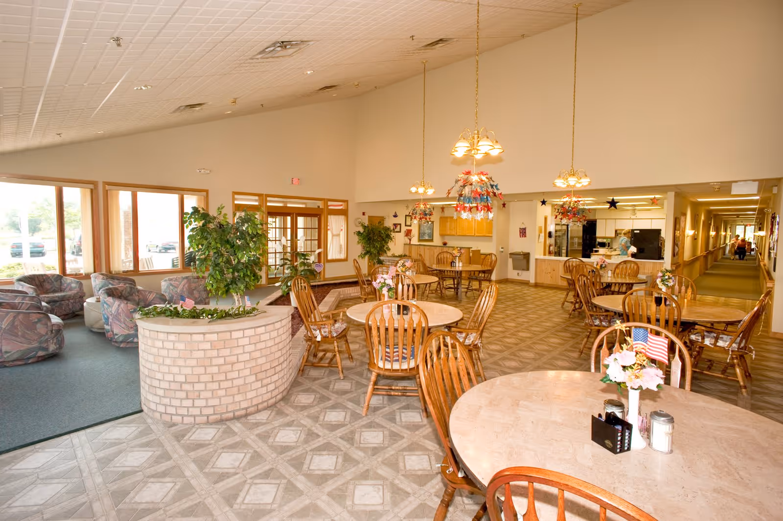 A spacious dining area in a senior living facility with round tables and wooden chairs. The tables have small flower arrangements with American flags. There are hanging light fixtures decorated with red, white, and blue star ornaments. To the left, there is a seating area with cushioned armchairs near large windows letting in natural light. The floor has a patterned tile design, and there are plants placed around the room. In the background, a kitchen area is visible with a person standing near the counter. A hallway extends from the dining area to the right.