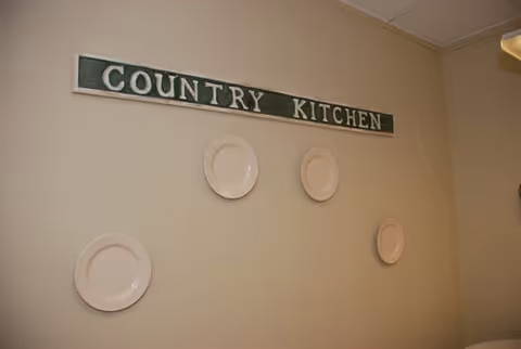 Wall with a green sign that reads 'COUNTRY KITCHEN' and five white decorative plates arranged around the sign.