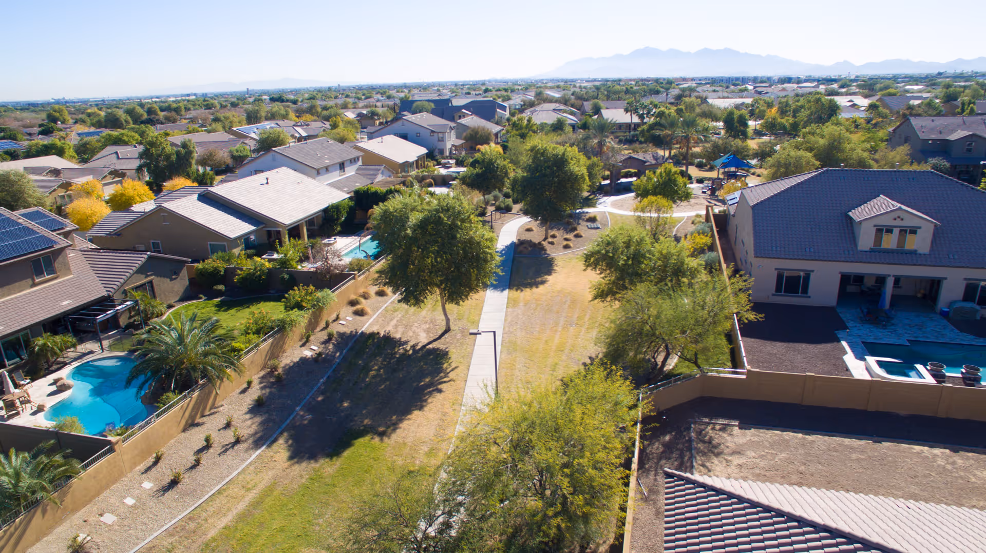 Aerial view of a residential neighborhood with houses, trees, and a walking path running through a grassy area. Some houses have swimming pools in their backyards, and mountains are visible in the distance under a clear sky.