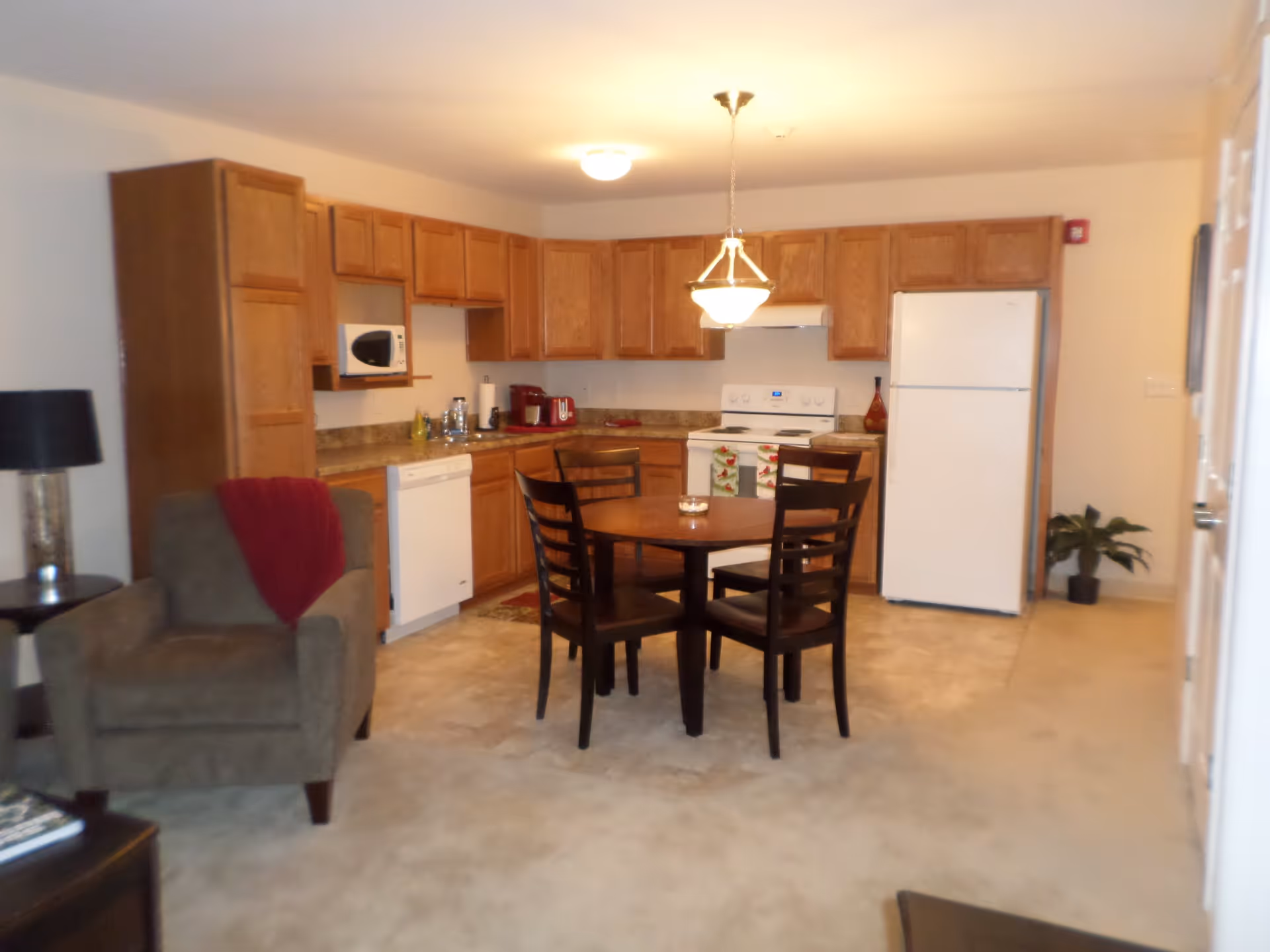 Interior view of a kitchen and dining area with wooden cabinets, a white refrigerator, stove, microwave, and dishwasher. A round wooden dining table with four chairs is in the center. To the left, there is a gray armchair with a red throw blanket and a side table with a lamp. The floor is carpeted and tiled near the kitchen area.