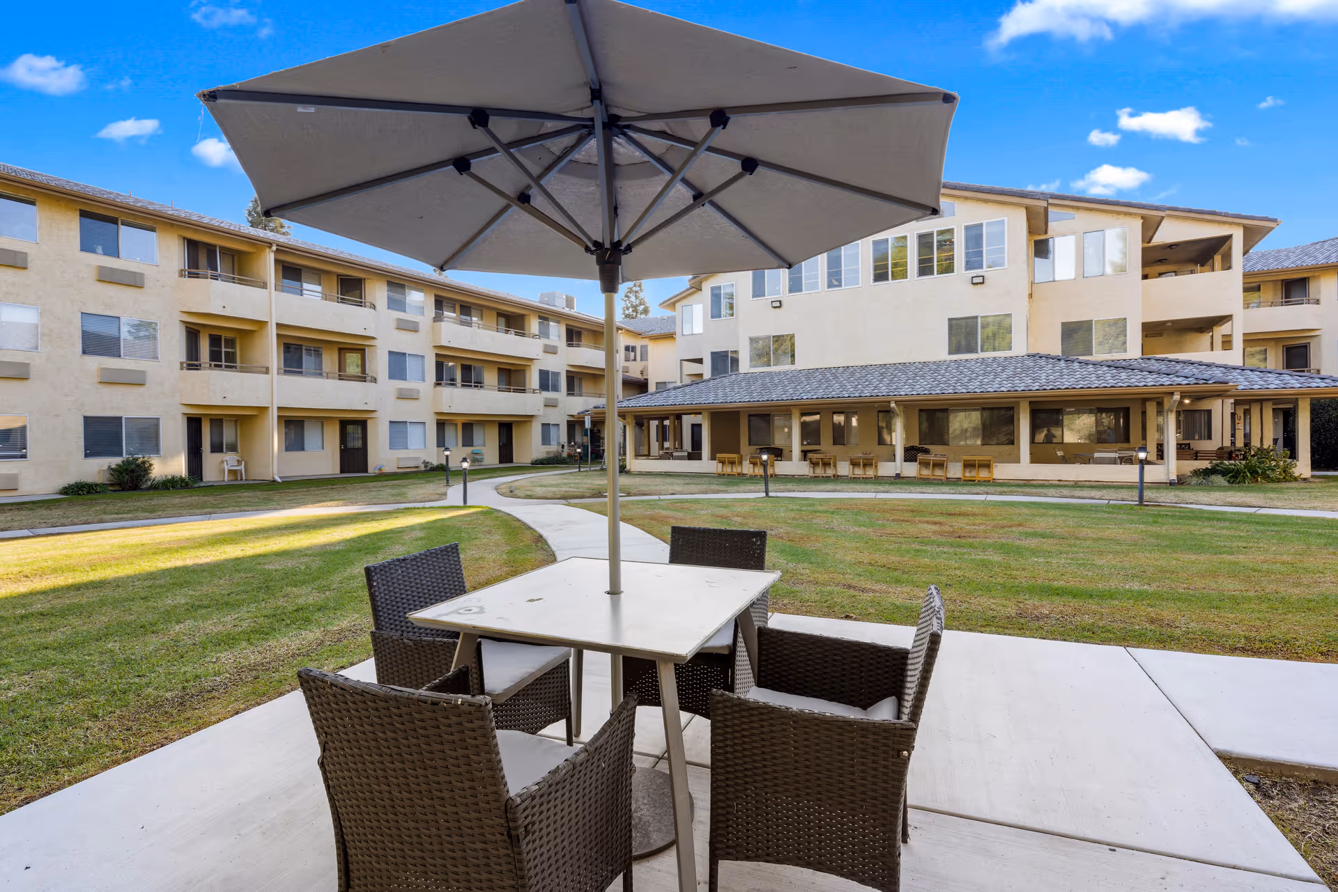 Outdoor patio area at Columbus Estates senior living community featuring a square table with four wicker chairs and a large umbrella, surrounded by a grassy courtyard and multi-story residential buildings under a blue sky with scattered clouds.