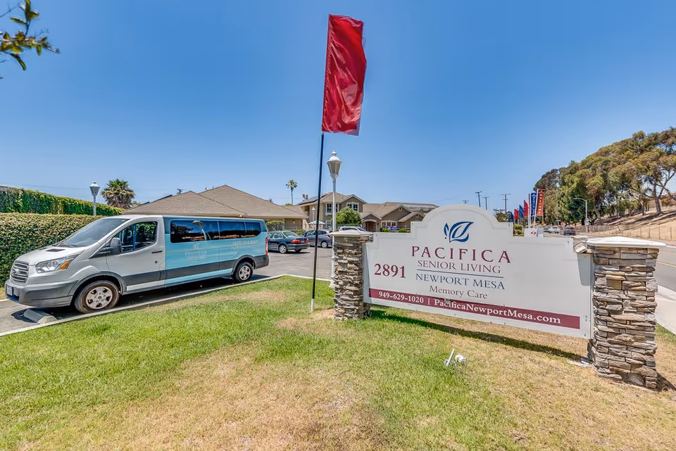 Outdoor view of the entrance to Pacifica Senior Living Newport Mesa Memory Care facility, showing a large sign with the facility's name, address, phone number, and website. A silver and blue shuttle van is parked nearby, with buildings and a clear blue sky in the background.