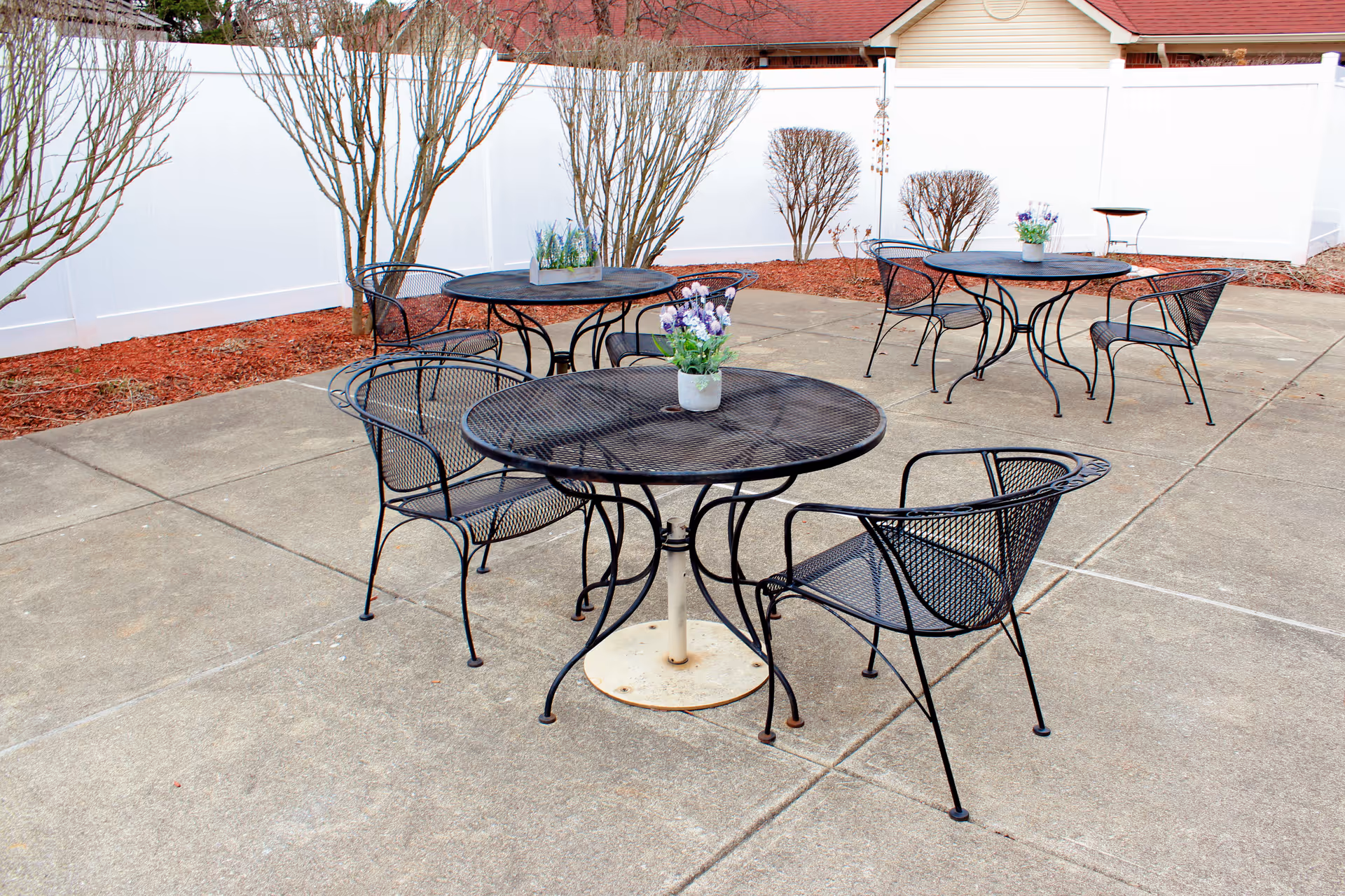 Outdoor patio area with several black metal tables and chairs arranged on a concrete surface. Each table has a small flower pot with purple and white flowers. The patio is enclosed by a white fence, and there are leafless trees and shrubs planted in mulch beds along the fence.
