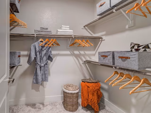 A neatly organized walk-in closet with wooden hangers on metal rods, gray fabric storage bins on shelves, a gray robe hanging on the left side, folded towels on the top shelf, a wicker laundry basket, and an orange throw blanket draped over a small stool.