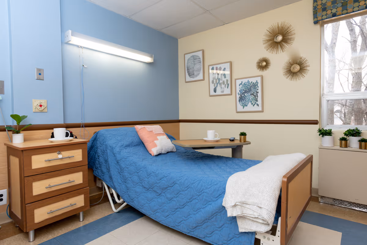 A nursing facility bedroom with a single bed covered in a blue quilt and a white blanket folded at the foot. There is a wooden nightstand with three drawers and a white cup on a saucer. A small table extends over the bed with a white cup and small potted plant on it. The walls are painted blue and beige, decorated with framed abstract art and gold sunburst wall decor. A window with a patterned valance lets in natural light, and several small potted plants sit on the windowsill.