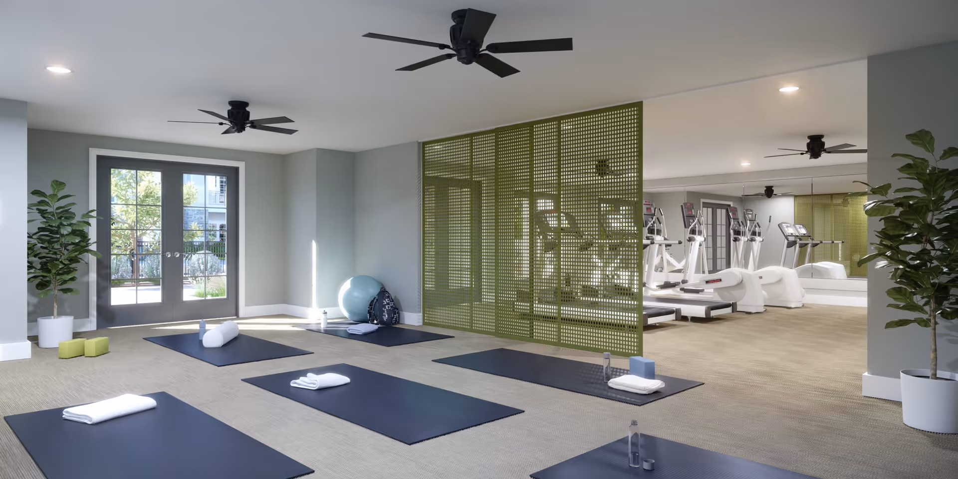A bright fitness room with exercise mats, towels, water bottles, and yoga blocks arranged on the floor. The room has large glass doors letting in natural light, two ceiling fans, potted plants, and a green perforated partition separating the yoga area from a gym area with treadmills and other exercise equipment reflected in a large mirror.