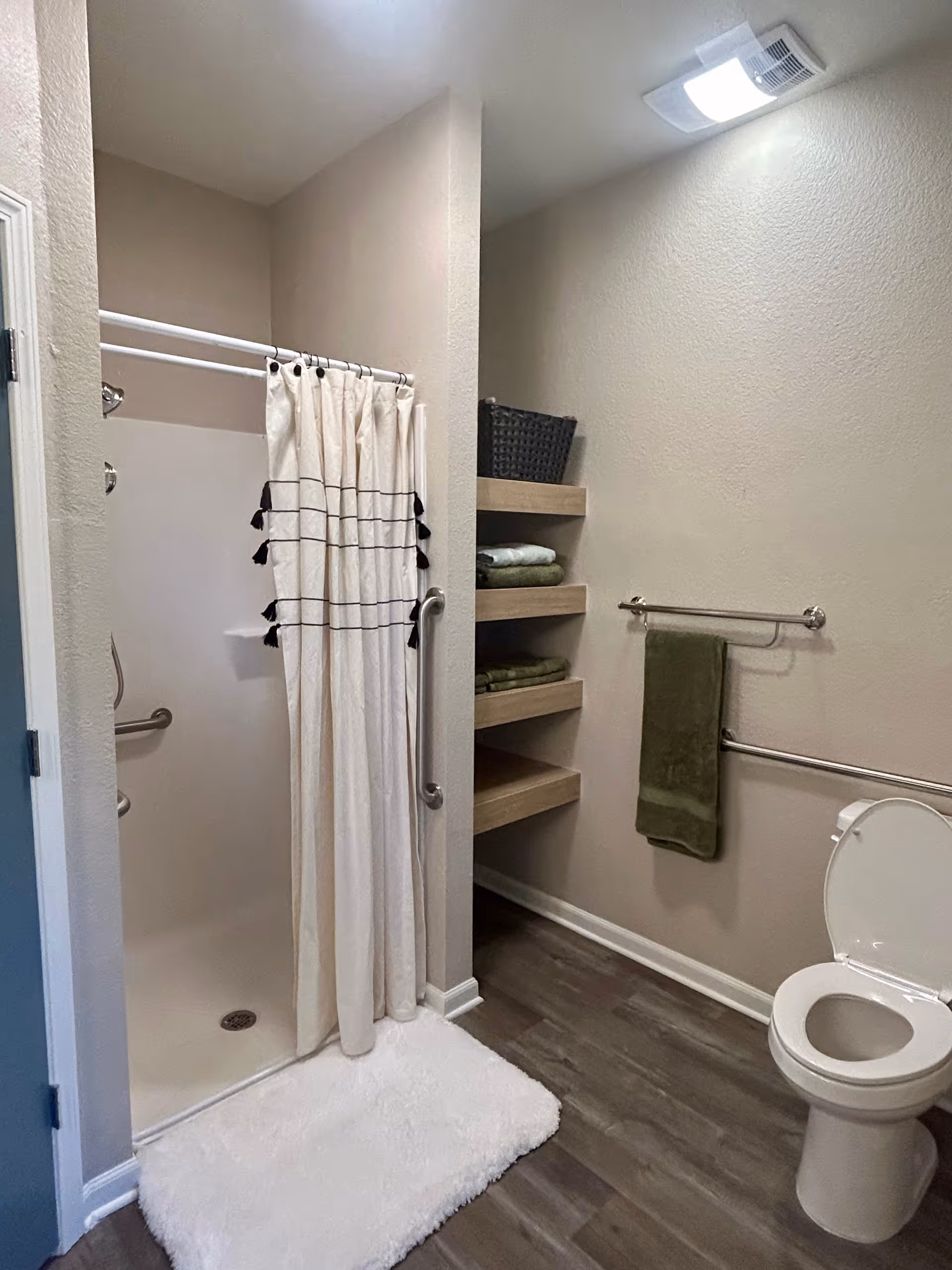 A bathroom with a walk-in shower featuring a white curtain with black tassels, a white fluffy bath mat, a toilet with the lid open, and built-in wooden shelves holding folded green towels and a black basket. The walls are beige and the floor is wood-style vinyl. There are two towel bars on the wall, one with a green towel hanging.