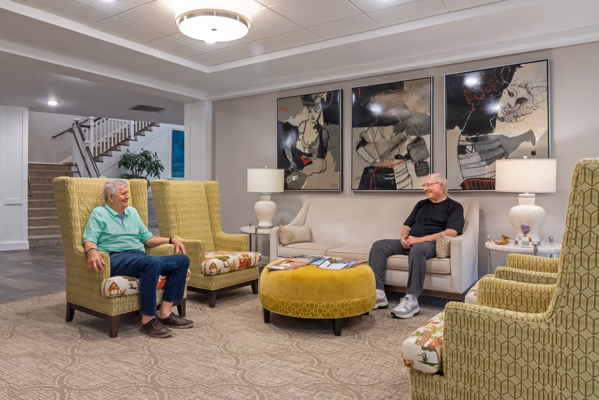 Two elderly men sitting and chatting in a well-lit senior living facility lounge area with patterned armchairs, a beige sofa, a round yellow ottoman, and three large artistic portraits on the wall behind the sofa.