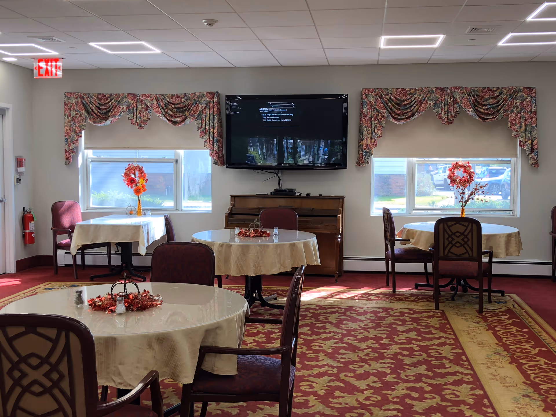 A dining room with round and square tables covered with beige tablecloths, each decorated with a centerpiece of flowers. The room has patterned red and gold carpet, floral valances over the windows, a wall-mounted flat screen TV above a piano, and several upholstered chairs around the tables. An exit sign and fire extinguisher are visible on the left wall.
