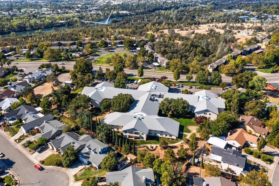 Aerial view of Hilltop Estates, a senior living community surrounded by trees and residential houses, with a river and a pedestrian bridge visible in the background.