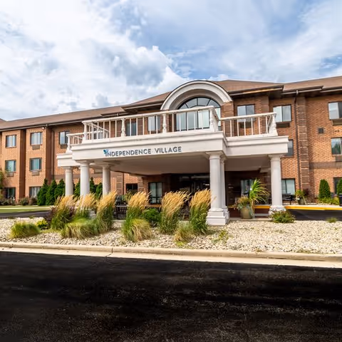 Front exterior view of Independence Village senior living facility showing a three-story brick building with a covered entrance supported by columns, landscaped with ornamental grasses and plants under a partly cloudy sky.