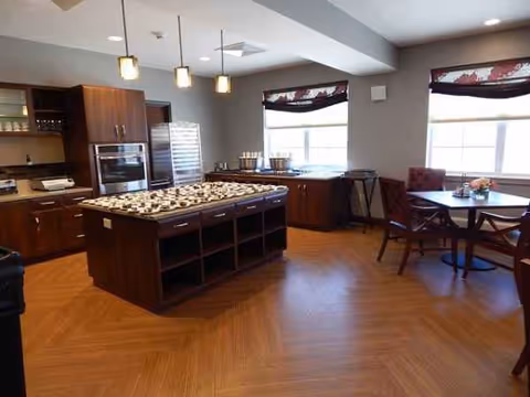 Interior communal kitchen with central island, oven, fridge, and dining table in a senior living facility.