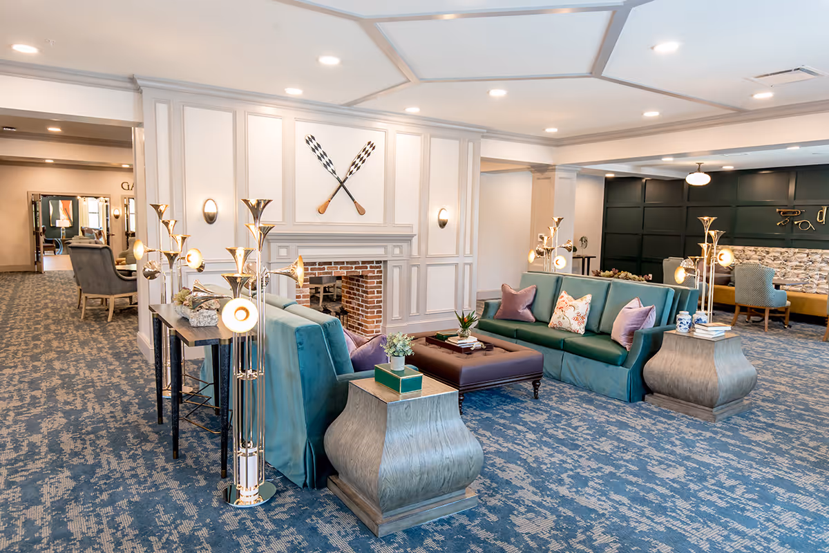 A spacious and elegant living room area in a senior living facility featuring two teal sofas with decorative pillows, a large ottoman coffee table, unique modern floor lamps, and a fireplace with crossed oars mounted above it. The room has a patterned blue carpet, white paneled walls, and recessed lighting in the ceiling. Additional seating and decor are visible in the background.