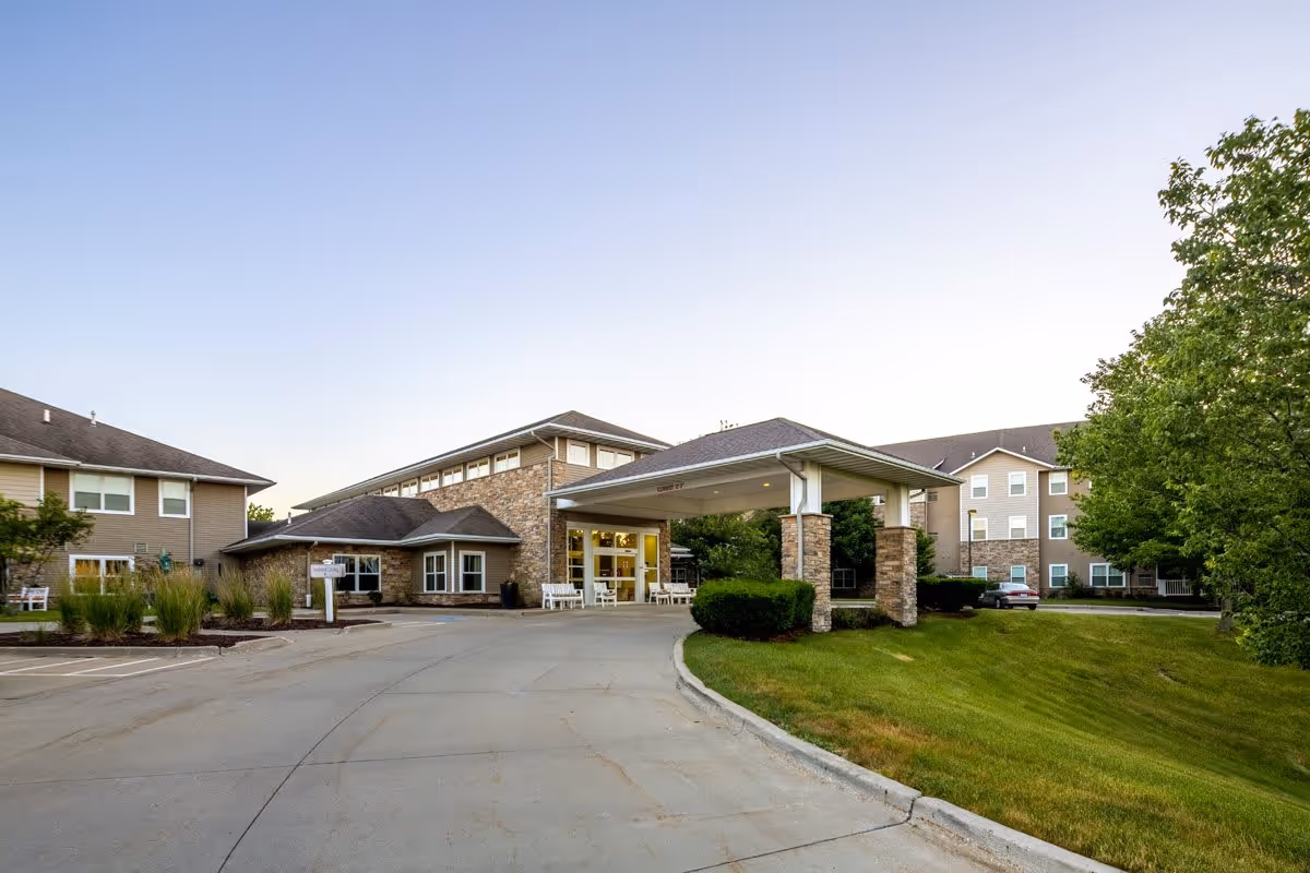 Exterior view of Independence Village of Waukee showing the entrance with a covered drop-off area supported by stone pillars, surrounded by well-maintained landscaping and multiple building wings under a clear sky.