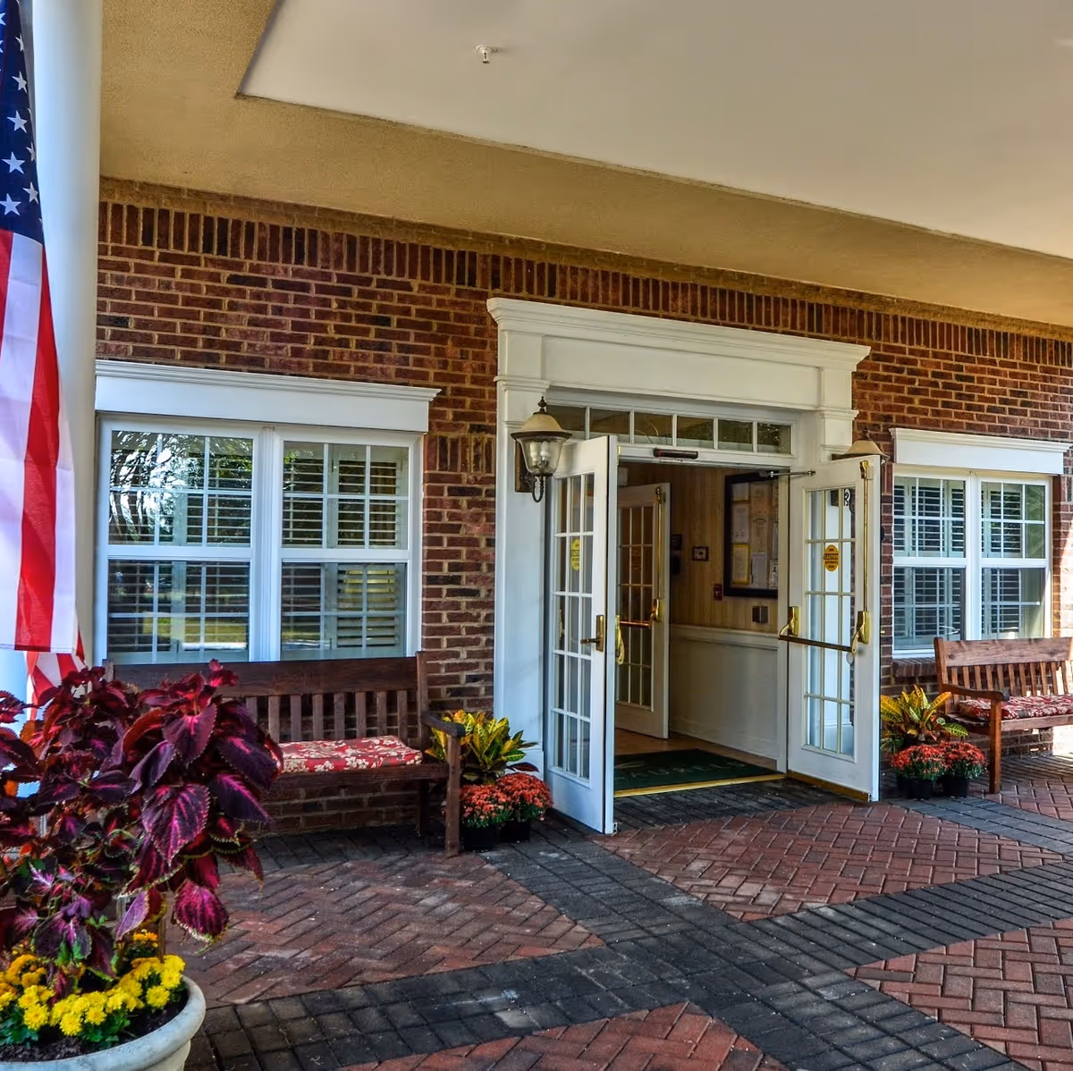 Entrance to a brick building with double glass doors open, flanked by two windows with white shutters. There are wooden benches with floral cushions on either side of the entrance, potted plants with colorful flowers, and an American flag on the left side.