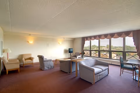 A spacious living room in a senior living facility with multiple armchairs and sofas arranged around a small table. There is a TV on a stand in the corner and large windows with curtains letting in natural light, overlooking an outdoor view.