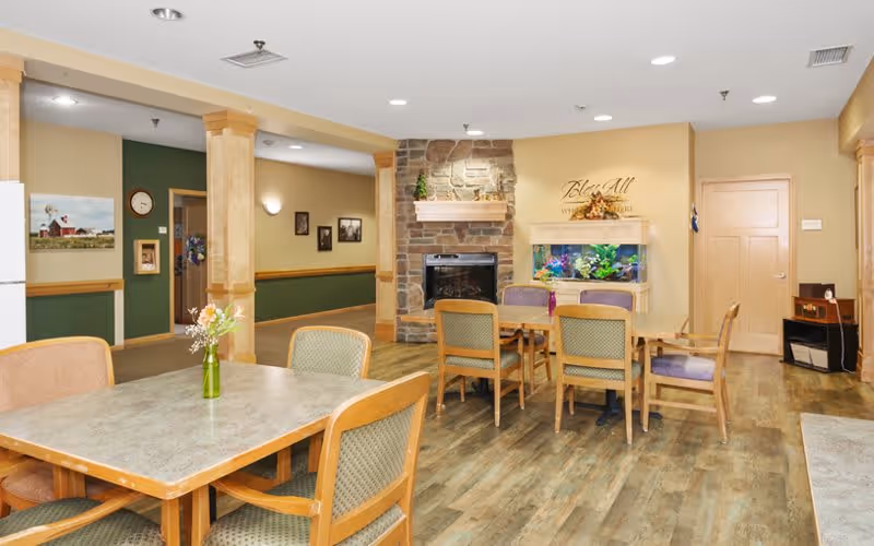 A cozy common area in an assisted living facility featuring wooden tables and chairs arranged on a wood-patterned floor. There is a stone fireplace in the background with a wooden mantel, above which is a decorative arrangement. Next to the fireplace is a large aquarium filled with colorful fish. The walls are painted in warm tones with some green accents, and framed pictures hang on the walls. The space is well-lit with ceiling lights.