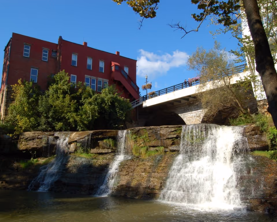Red brick building and stone bridge above a cascading waterfall with trees and a clear blue sky.