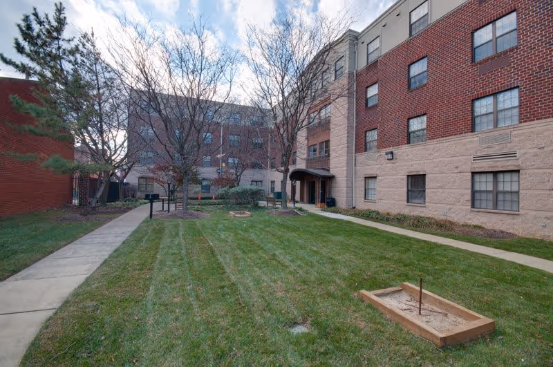 Outdoor courtyard area of a senior living facility with green grass, leafless trees, a horseshoe pit, and a paved walkway leading to the building entrance. The building has a brick and stone exterior with multiple windows.
