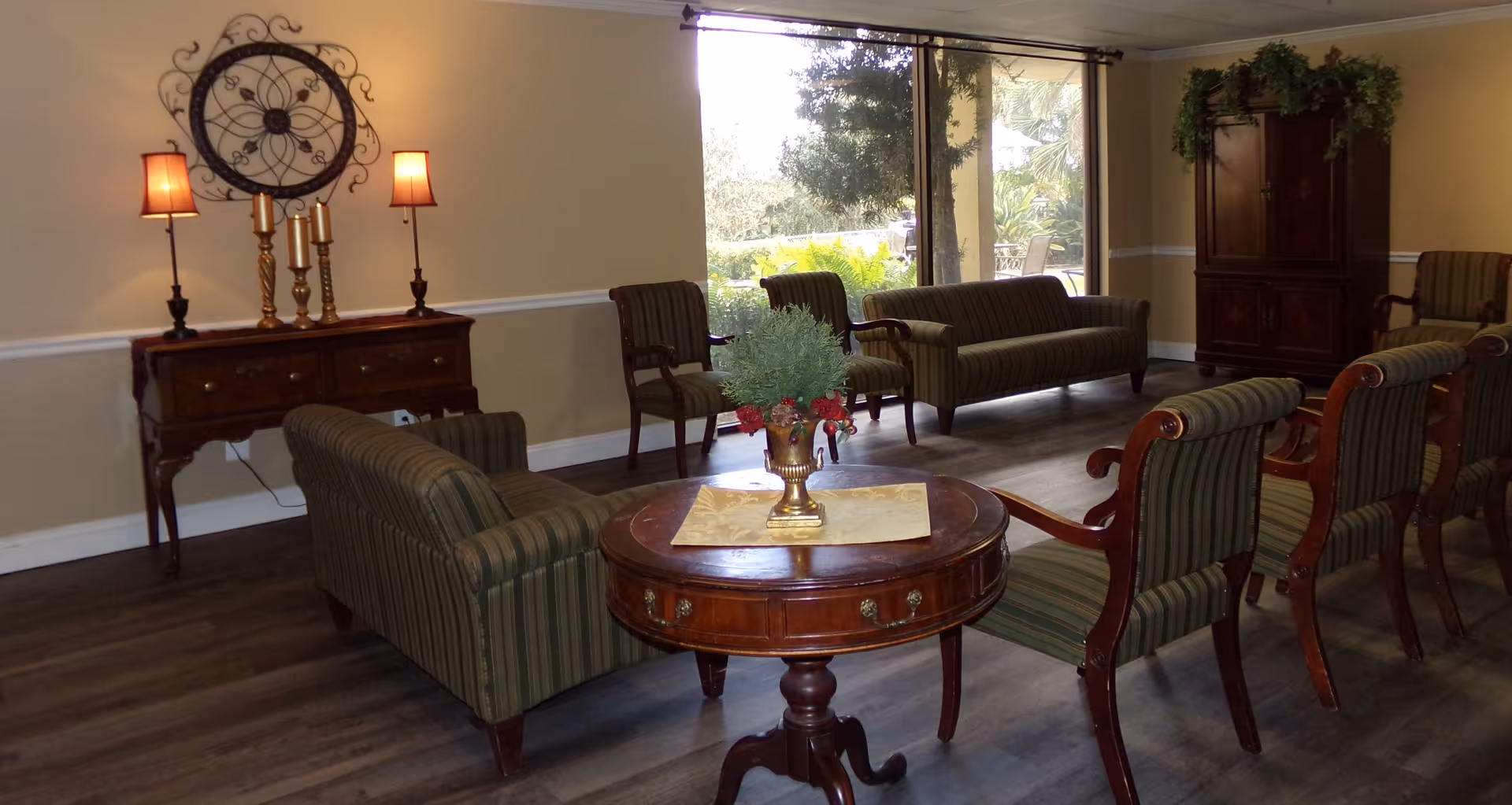 A cozy living room area with striped upholstered sofas and chairs arranged around a round wooden table with a decorative plant centerpiece. There is a wooden sideboard with two lamps and a large decorative wall clock above it. A large window lets in natural light and shows greenery outside. The room has wooden flooring and neutral-colored walls.