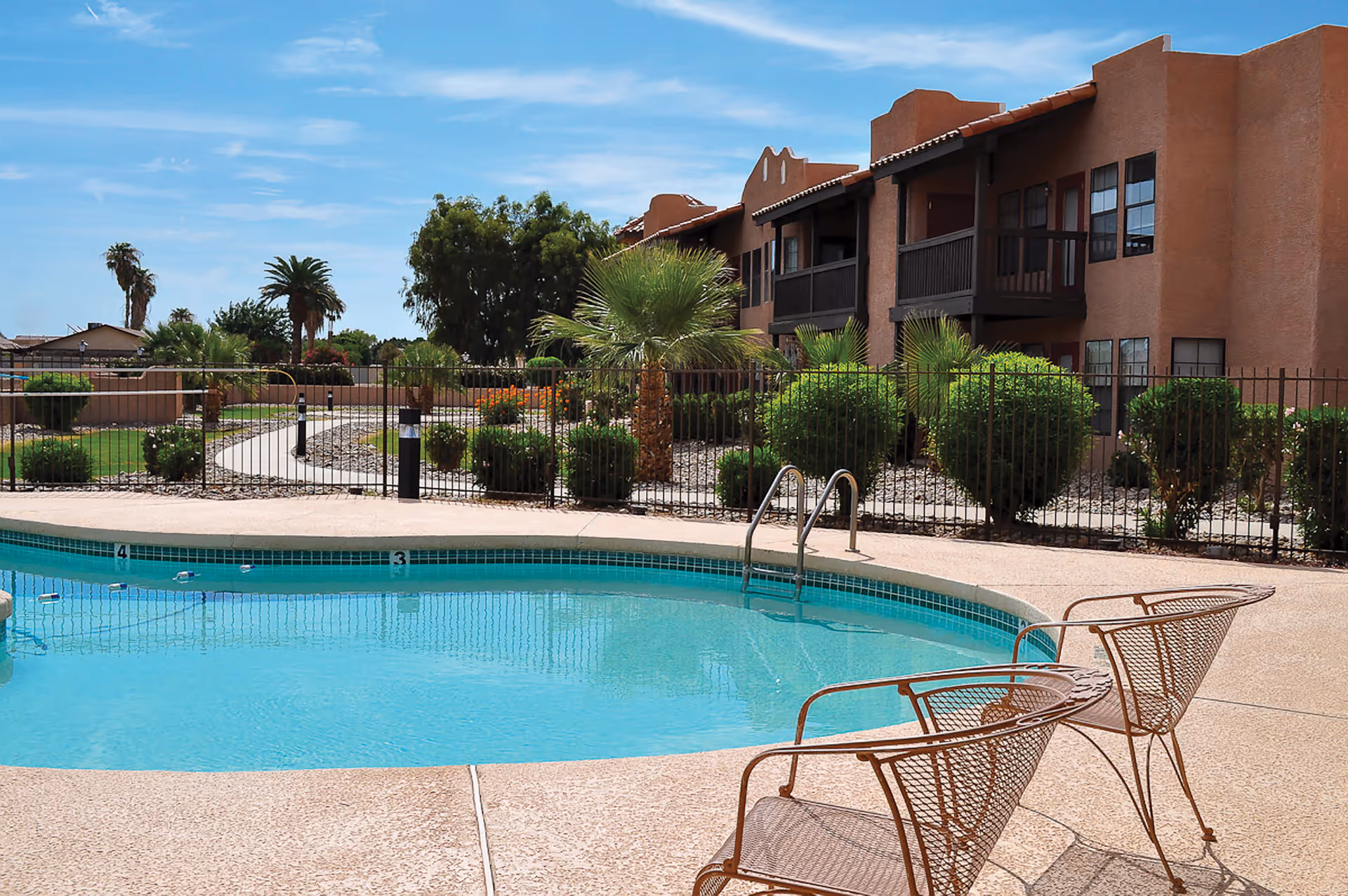 Outdoor swimming pool with clear blue water surrounded by a concrete deck. Two metal mesh chairs are positioned near the pool. In the background, there is a fenced garden area with palm trees, bushes, and a walking path. A two-story building with balconies and windows is visible behind the garden under a blue sky with some clouds.