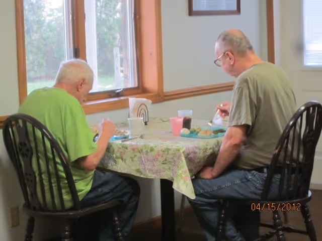 Two older men sit at a small round table by a window eating a meal.