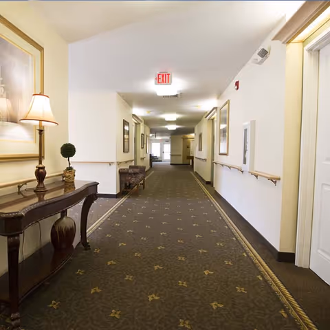 A well-lit hallway in a senior living facility with patterned carpet, handrails on both sides, framed artwork on the walls, a wooden console table with a lamp and decorative plant, and an exit sign at the far end.