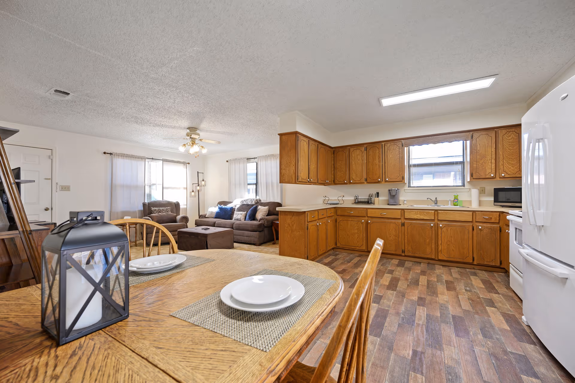 Interior view of a senior living facility showing a combined kitchen and living room area. The kitchen has wooden cabinets, a white refrigerator, stove, microwave, and a window above the sink. The living room area includes a brown sofa, armchair, ottoman, and a floor lamp near two windows with white curtains. A wooden dining table with two place settings and a decorative lantern is in the foreground.