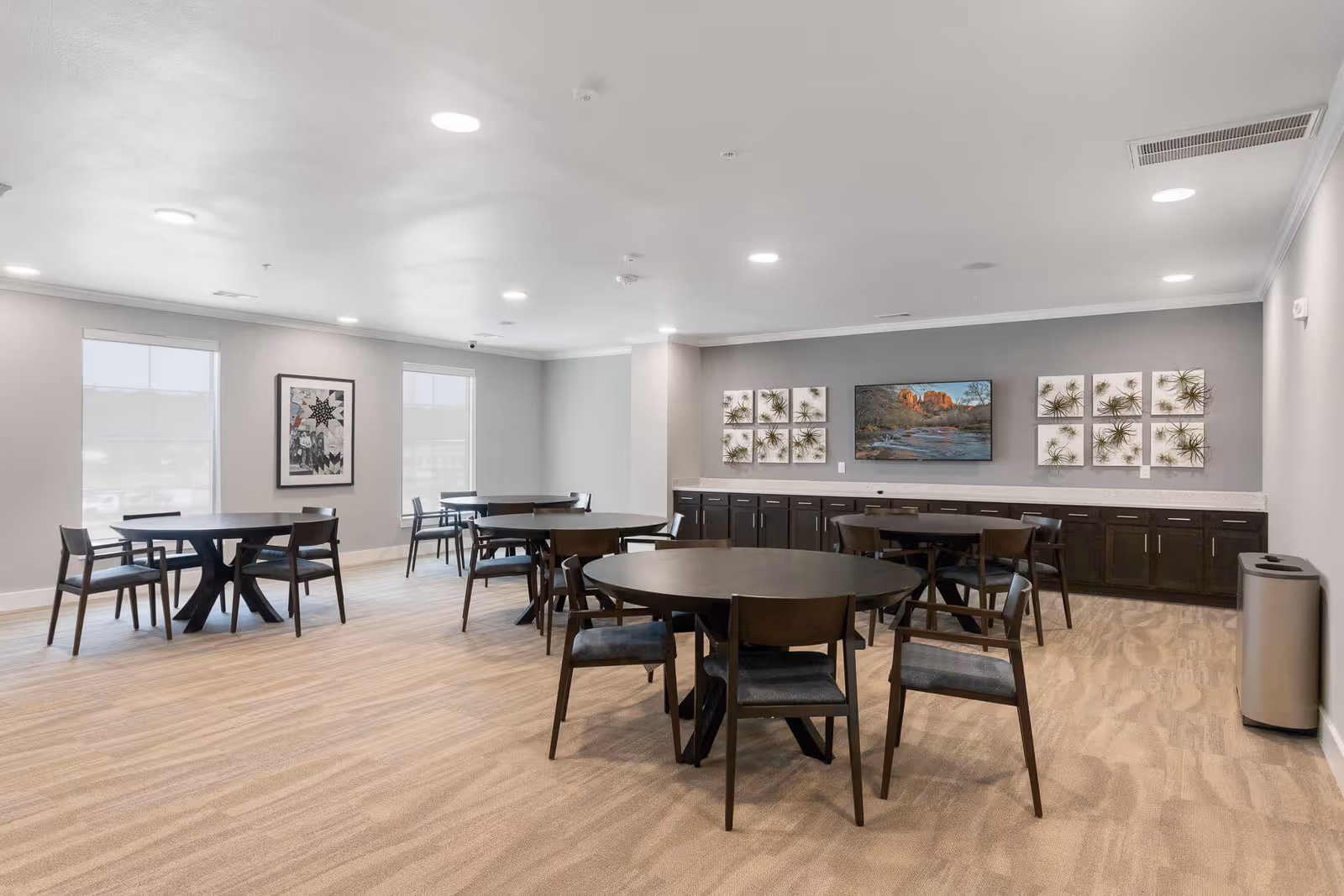 A spacious dining room with several round dark wood tables and matching chairs arranged on a light carpeted floor. The walls are painted light gray with two large windows on one side and decorative artwork on the opposite wall above a long counter with dark cabinetry. The room is well-lit with recessed ceiling lights.