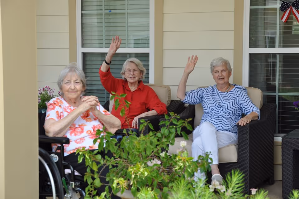 Three elderly women sitting on a porch in comfortable chairs. Two of them are waving and smiling, while the third woman in a wheelchair is sitting with her hands clasped. There are green plants in the foreground and windows with blinds behind them. An American flag decoration is visible on the right side.