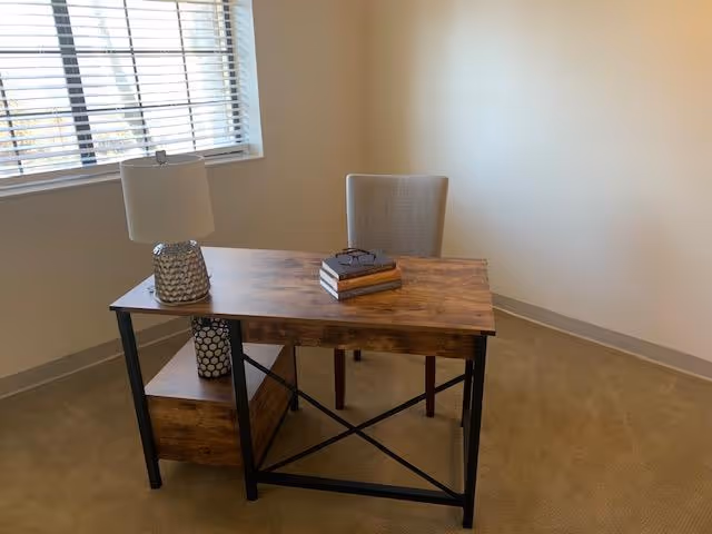 A small wooden desk with black metal legs placed in a corner of a room with beige walls and carpet. On the desk are a table lamp with a textured base, a stack of three books, and a pair of eyeglasses. Behind the desk is a beige upholstered chair. A window with white blinds is on the left wall.