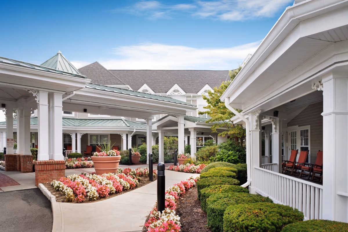 Covered porte-cochere entrance of a senior living building with colorful flower beds and a porch with red rocking chairs.